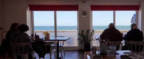 This image shows the interior of Cafe 1, located in Devon, England, United Kingdom, with people seated at tables near large windows that offer a clear view of the sea. The photograph is taken in the afternoon during early June, indicating a late spring season. The type of photograph is an indoor scene that combines elements of still life and social documentary. The scene captures people enjoying drinks and food inside the cafe, with the blue sea visible outside, suggesting a scenic coastal location. The decor includes plants and a vase of flowers on the tables, while natural light streams in through the windows, highlighting the relaxed atmosphere and the presence of people inside the cafe.